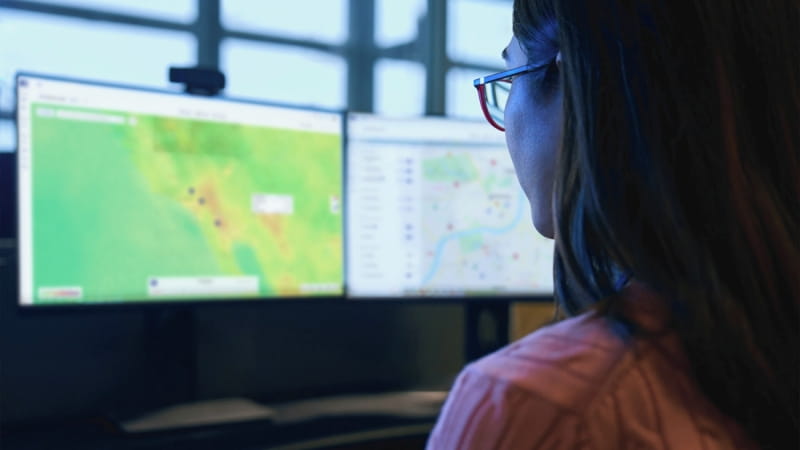 Person vor mehreren Bildschirmen mit Karten- und Analysedaten einer Sicherheits- und Risikomanagementplattform. Die Monitore zeigen geografische Informationen und Echtzeitdaten zur Überwachung von Risiken und Ereignissen.